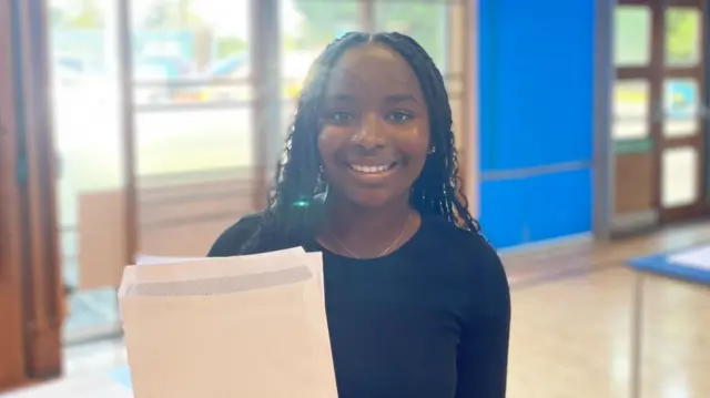 A girl in a black top stands in a school gymnasium, she holds a white opened envelope and is smiling