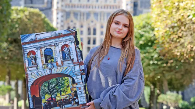 A girl with blonde straight hair and a grey sweartshirt holds a piece of art she made and stands outside Brighton College