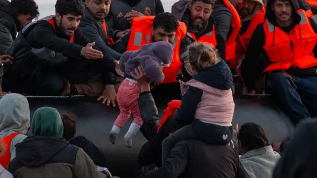 Two young children being passed to a man on a black dingy ahead of an attempt to cross the Channel. Several people in orange life jackets are on the boat, while others are approaching it