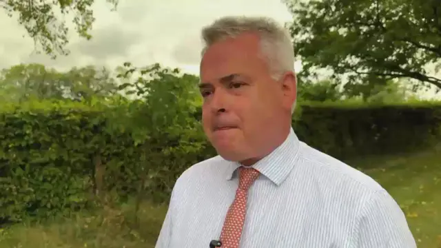 Close up image of Tim Loughton wearing shirt and tie in front of a hedge