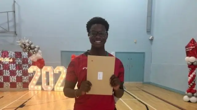 Holding up a brown envelope, Gabriel, wearing galsses and with a big beaming smile, holds up his results.