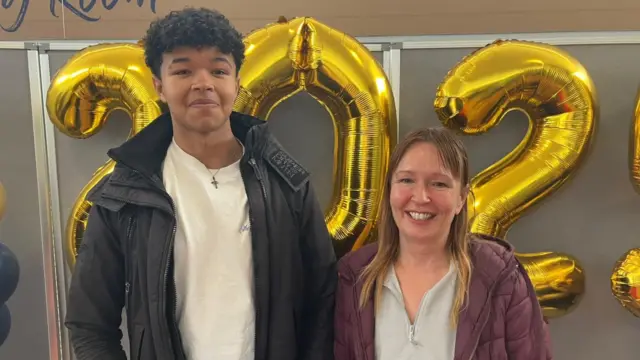 Noah stands next to his mum in front of some golden numbered balloons. They are both wearing coats and smiling at the camera