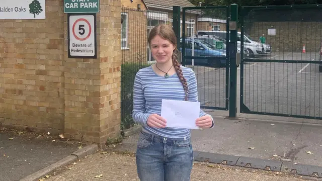 Betty wears a stripey blue jumper, jeans and holds an envelope containing her results