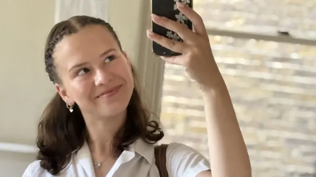 A girl with brown hair tied half back takes a mirror selfie, she wears a white tshirt and is standing in front of a cream wall and window