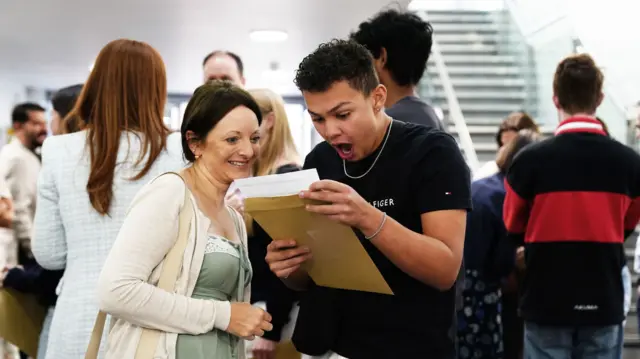 A boy in a black t shirt looks at an envelope of results with a shocked face while a woman looks over