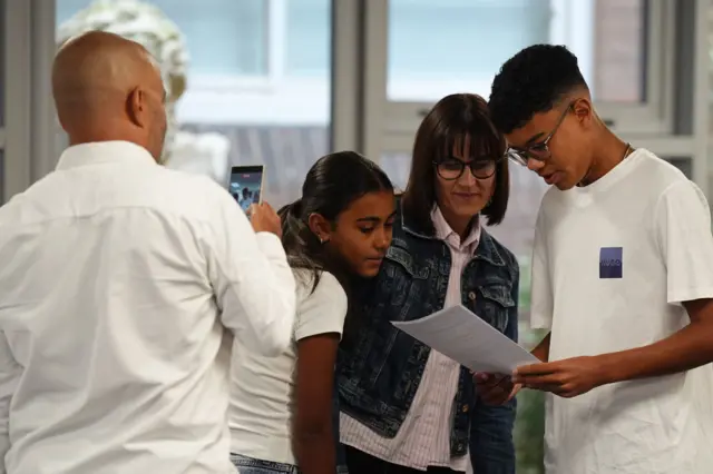 Boy reads results off white paper while woman and girl peer over to look at paper. A man stands back taking a picture of them on his phone