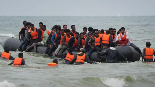 A group of men wearing life jackets sit in a small inflatable boat on a body of water.