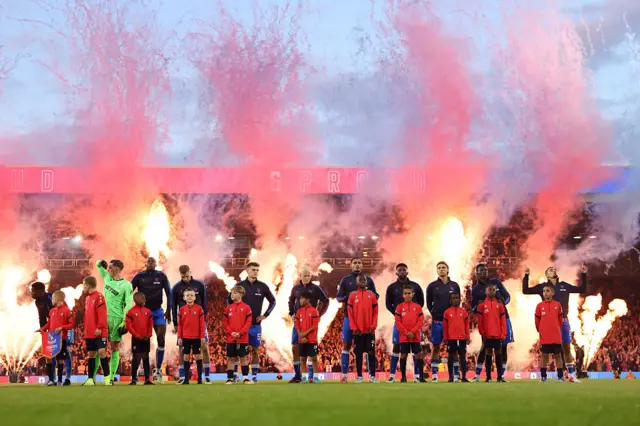 The players line up pre kick off with flares in the background