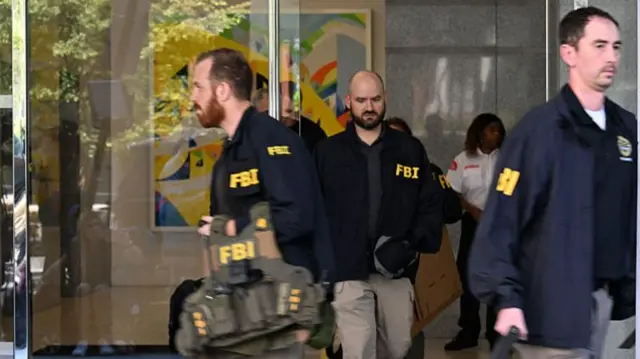 FBI agents walk out of the glass doors of Bolton's office holding bags and wearing jackets clearly marked with 'FBI'