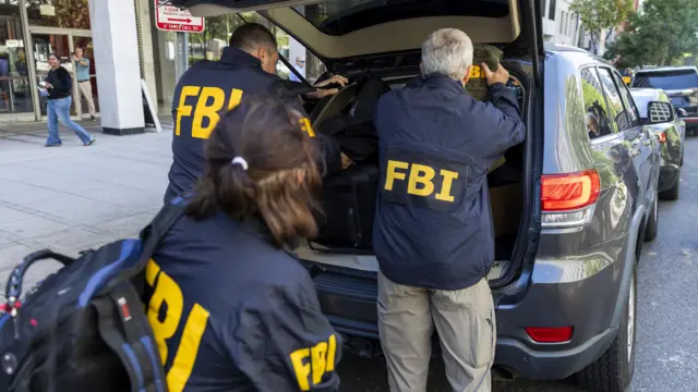 Three people wearing jackets emblazoned with FBI initials load items into the back of an SUV