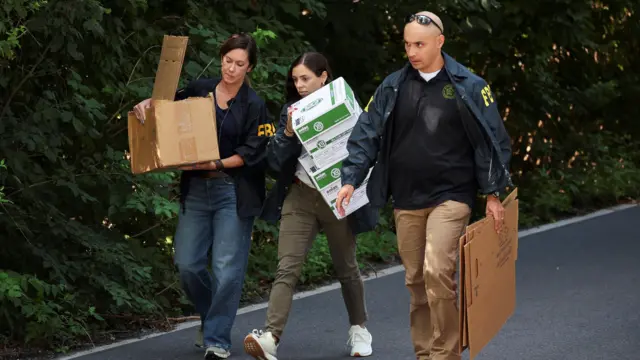 FBI officials carry cardboard boxes, two women in casual wear with FBI jackets carrying boxes to the left and one man with folded boxes in his left hand to the right. They're walking along a road, shrubbery to the side of it