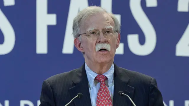 Close up of John Bolton in pin-striped black suit, striped light-blue shirt and patterned red tie. He's delivering a speech in front of a blue wall with white writings
