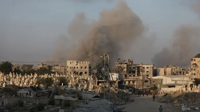Black and grey smoke billows over the ruins of damaged homes and apartment buildings in Gaza City following an air strike. A small group of people can be seen walking towards the buildings in the bottom right of the photo