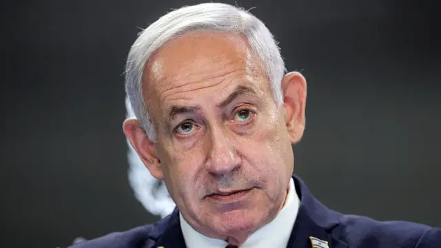 Close up of Benjamin Netanyahu in a dark blue suit, a Israeli flag pin on his left lapel, a black background behind him