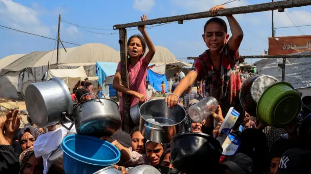 Children and women clamour for food at a charity kitchen in Khan Younis. They're holding out plastic and metal pans, pots and other containers. One girl in a pink top is holding on to a metal pole