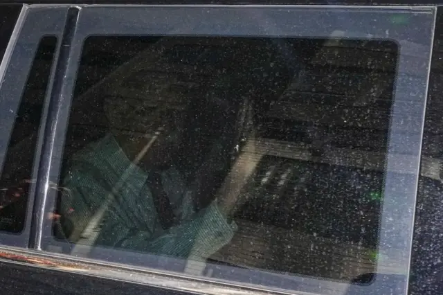 John Bolton himself, barely visible through the dusty car window, is pictured waving from a car leaving the building that houses his office
