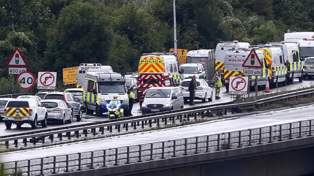Emergency service vehicles crowd onto a A-road amongst discarded cars.