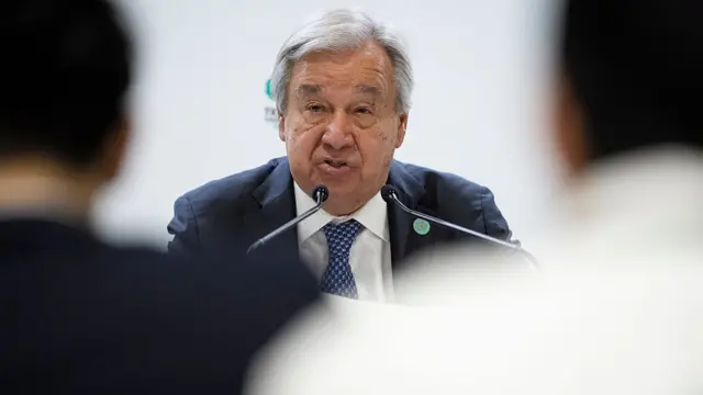 Close up of UN secretary general Antonio Guterres sitting down in a dark suit while speaking into two thin, black mics