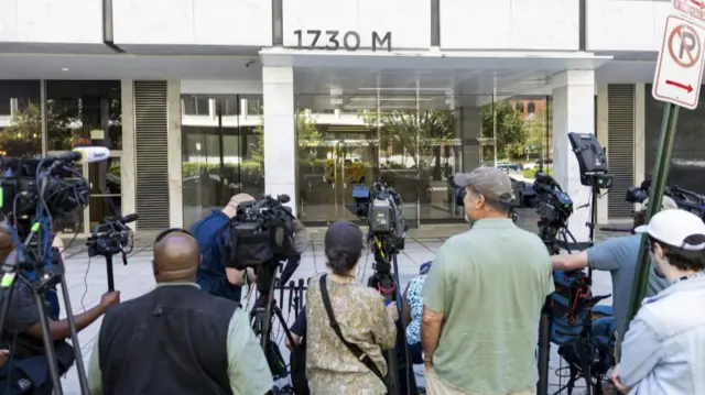 Members of the media holding large cameras outside a large building with glass doors, with '1730M' written above them