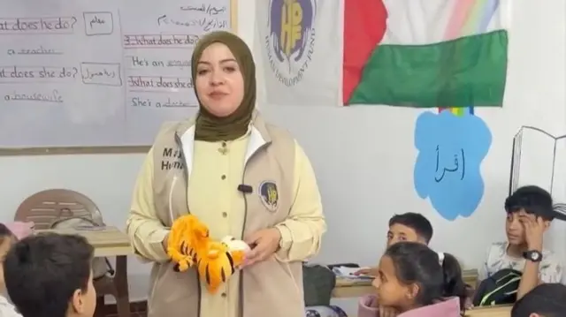 A woman holds a puppet as she stands in front of a whiteboard in a classroom.