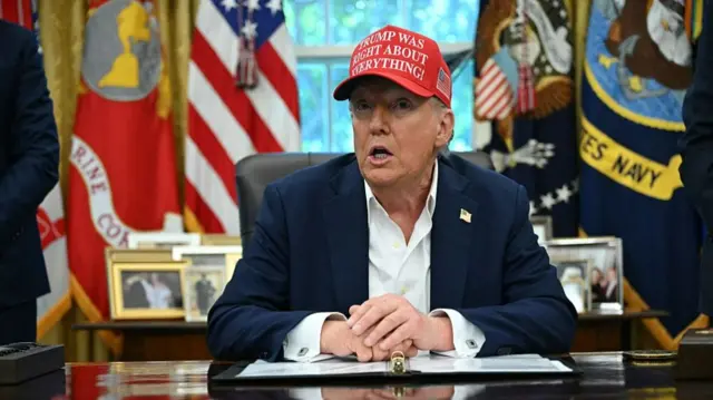 Trump, wearing a dark jacket and white shirt, sits in the Oval Office at his desk. He is also wearing a red hat that reads "Trump was right about everything"