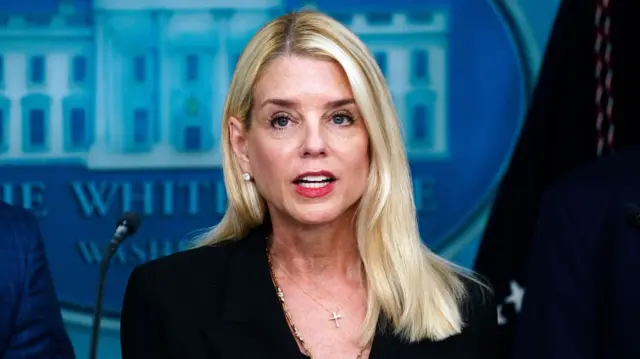 Pam Bondi in a black blazer addresses the White House press pool in the building's press room, the White House logo on a wall behind her