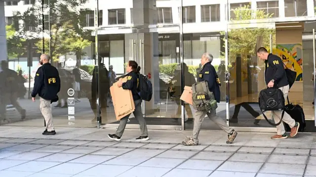 FBI agents wearing their blue jackets and holding boxes and cartons walk past large glass windows