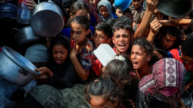 Large group of children holding metal pans and pots screaming and shouting for food at a charity kitchen in Khan Younis