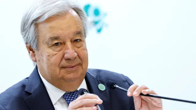 Antonio Guterres in a navy blue suit, white shirt and blue patterned tie - he is tilting a microphone towards himself