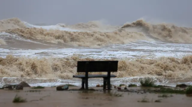 Strong waves head towards beachfront, a blurred bench facing the sea in the foreground