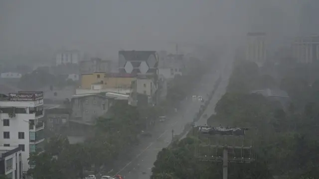 A grey picture with low clouds and rain. A road is at the centre of the image with trees on the right-hand side and buildings clustered on the left. Two cars with their headlights on are travelling down the road