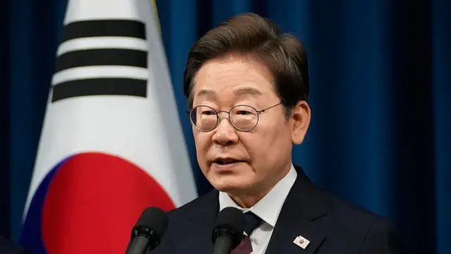 An elderly man with glasses stands in front of a microphone wearing a dark suit, a white shirt and a maroon and white tie. On his lapel there is a pin of South Korea.