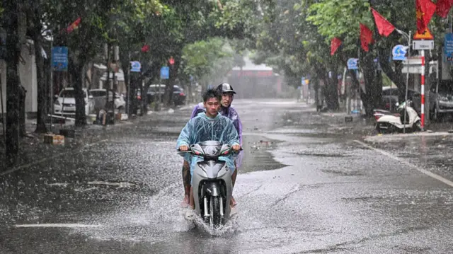 Two men on a motorbike wear waterproof ponchos as they travel towards the camera - water sits on top of the road