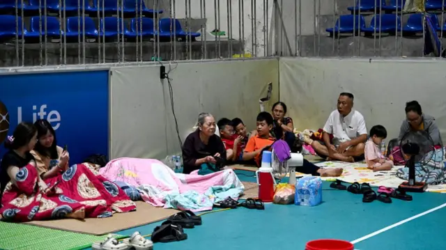 A mixture of young and old people sitting on mats in an indoor court. They are surrounded by sandals, and some of them are covered in blankets.