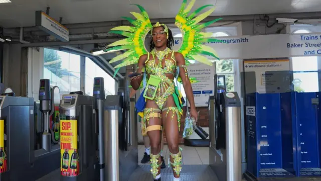 A woman in a green outfit with large glittery wings and feathers walking through a Tube barrier.