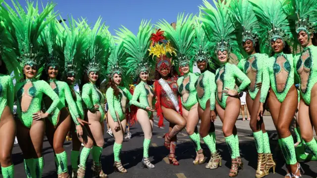 Women in green outfits and head dresses pose in a line, with a woman in red in the middle with a red headdress