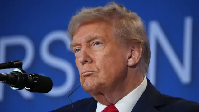 An elderly man purses his lips. He is sitting in front of a camera and he is wearing a dark suit, with a white shirt and a red tie. There is a small american flag pin on the front of his blazer.