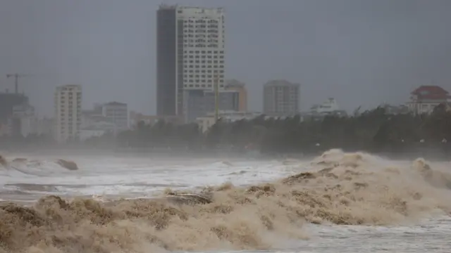A landscape image of waves crashing against a shoreline. The sky is grey and some high-rise buildings can be seen in the background