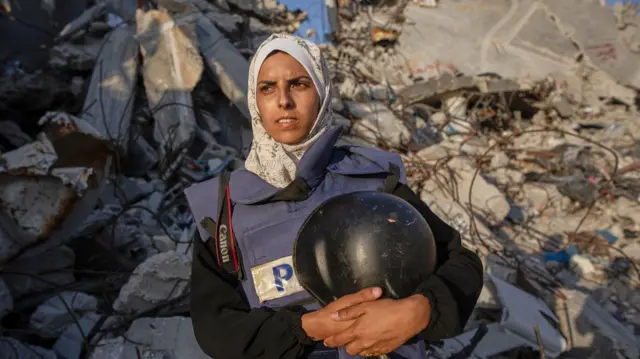 Mariam Dagga stands squinting into the sun wearing a press jacket and holding a helmet. Piles of rubble are behind her