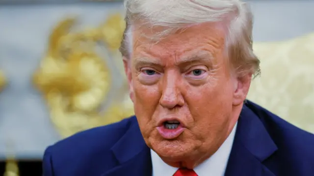 A close up photograph of Donald Trump's face while he speaks in the Oval Office, he wears a navy suit and red tie