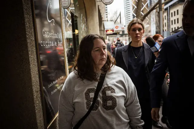 A woman in a frey sweater with the numbers "86" on its front walks down a busy street, flanked by people in suits and a microphone