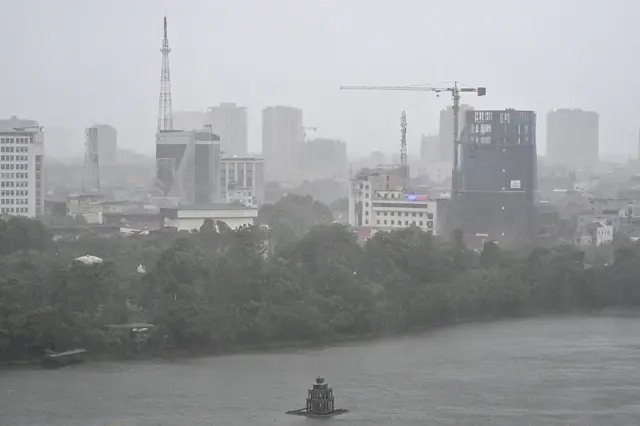 A view of tall buildings amid heavy rains and wind in Vinh city, Nghe. A row of trees buffers the urban area and the river.