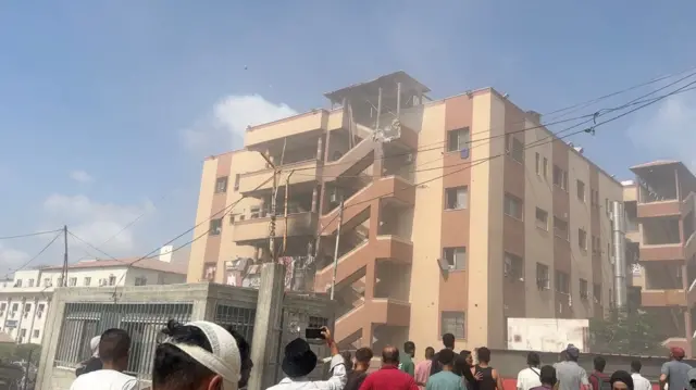 A landscape shot of a building which has been partly damaged. People stand around looking on - one takes a photo. A man with a bandage on his head is in the foreground