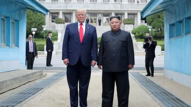 President Donald Trump and North Korean leader Kim Jong Un pose at a military demarcation line at the DMZ) in 2019. Both face staright forward. A photographer is seen taking their picture in the background.