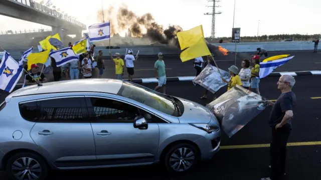 Protesters stand in front of cars on a main road with smoke in the background