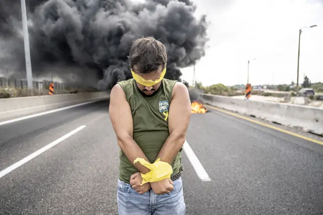 A man poses in the middle of the street, with his hands bound in yellow and his eyes covered with yellow tape.