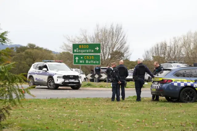 Police at the scene of a shooting in the high country of Porepunkah in Victoria. There are police cars in the background, and a road sign pointing to nearby towns.