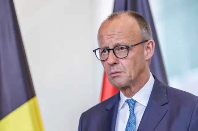 German Chancellor Freidrich Merz is looking off camera with a serious expression at a press conference with flags in the background. He is wearing a suit with a blue tie and glasses.
