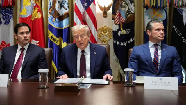 Donald Trump in suit say behind large desk, Marco Rubio in suit on the left and Pete Hegseth also in a suit on the right. US flags behind them.