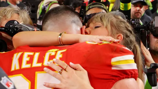 Taylor Swift hugs Kelce in a red jersey while crowds gather around